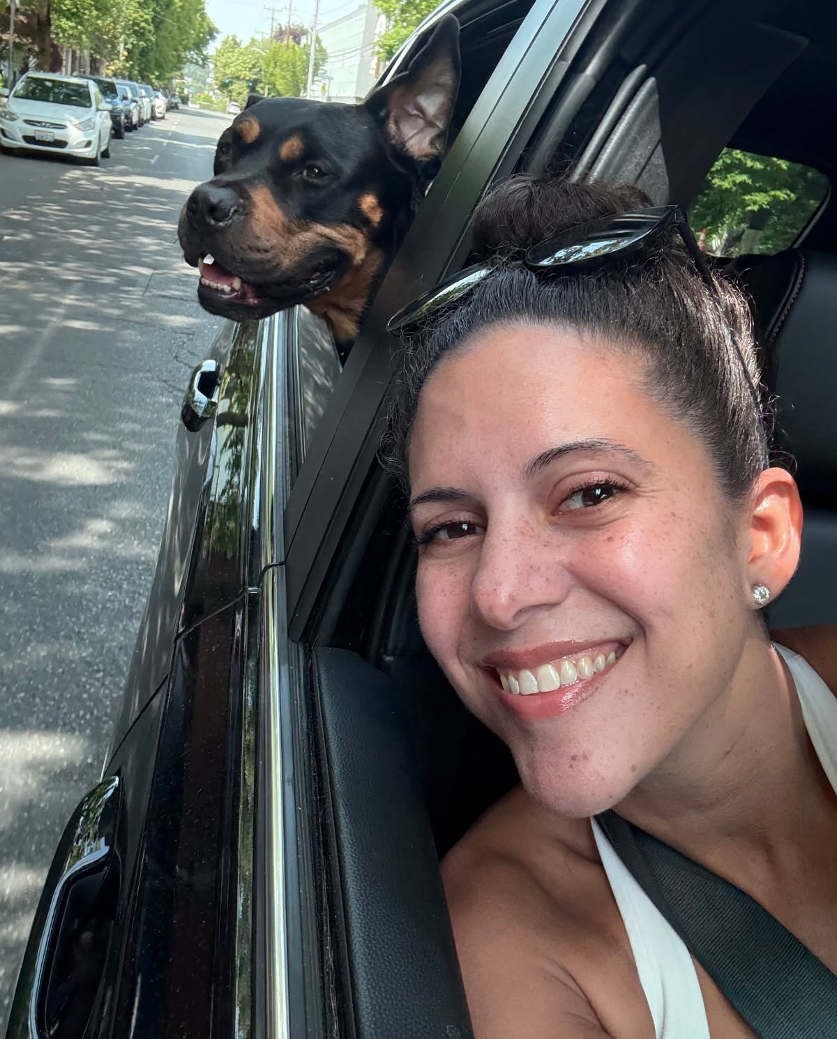 A smiling woman in a car with a happy Rottweiler looking out the window