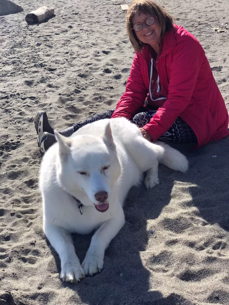 Adopter in a red jacket sitting on the beach with her happy white husky