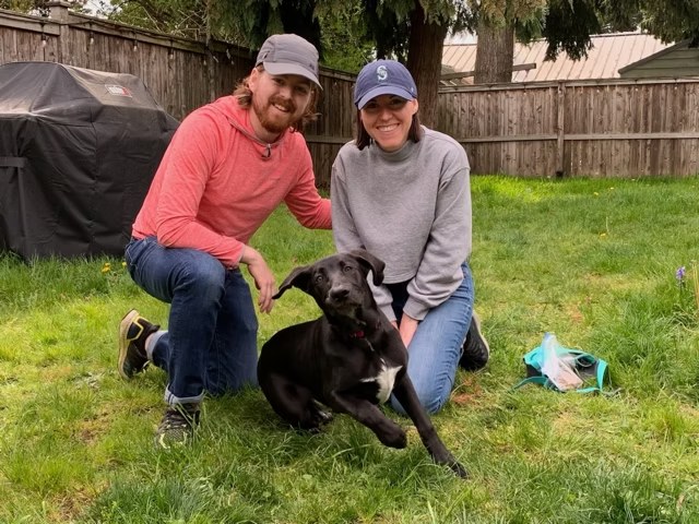 Couple kneeling in a green backyard beside their newly adopted black Lab mix